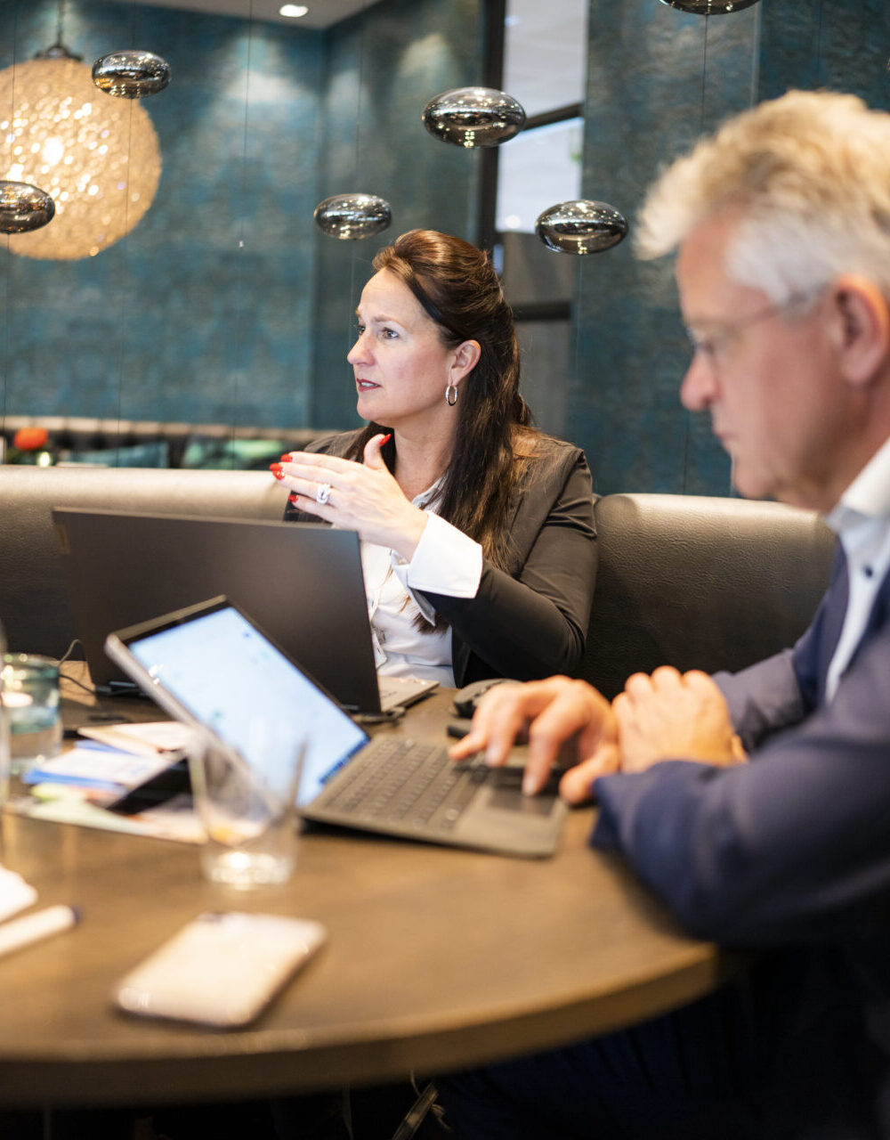 Een vrouw en een man zitten aan een ronde tafel met laptops en papieren voor hen.