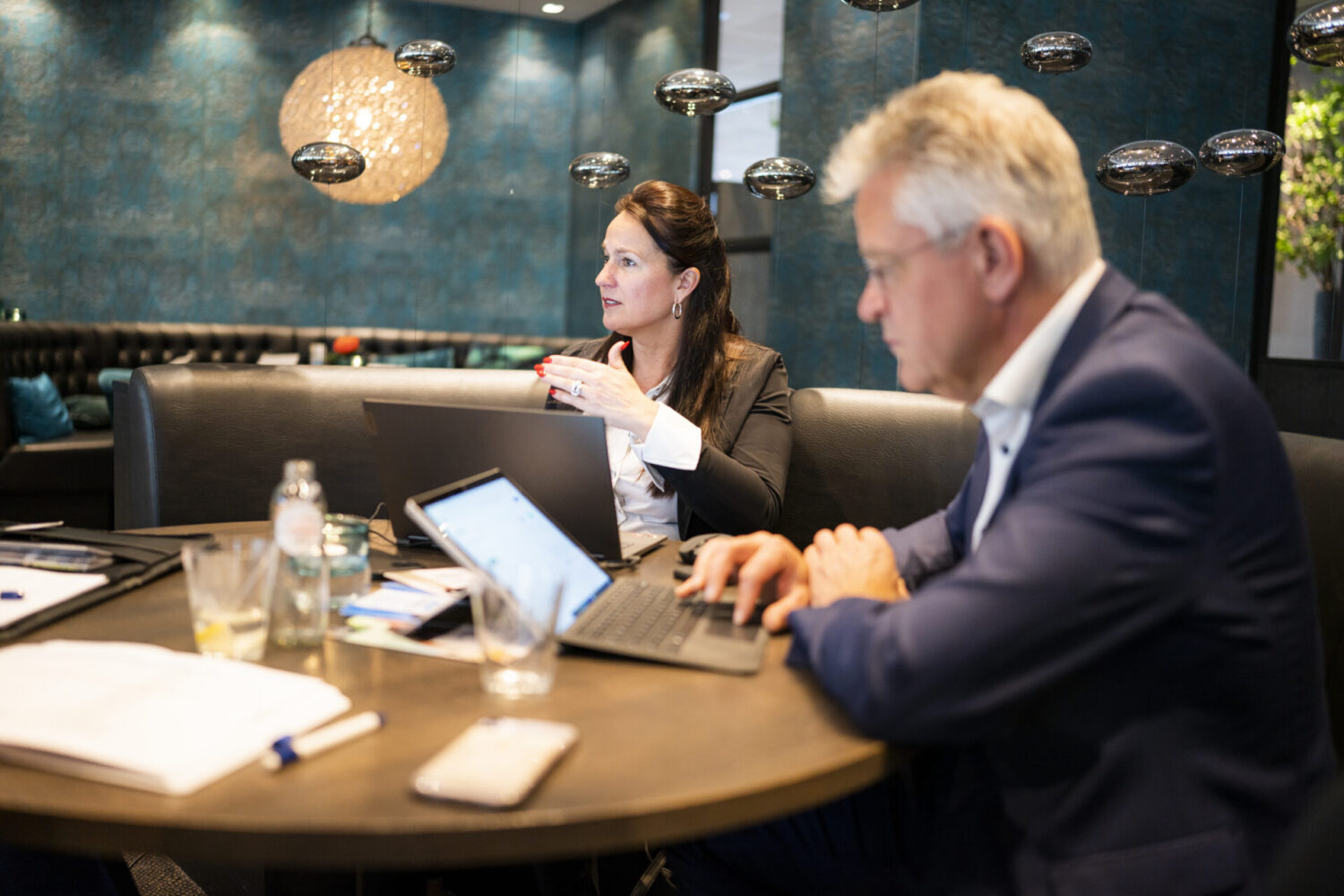 Een vrouw en een man zitten aan een ronde tafel met laptops en papieren voor hen.