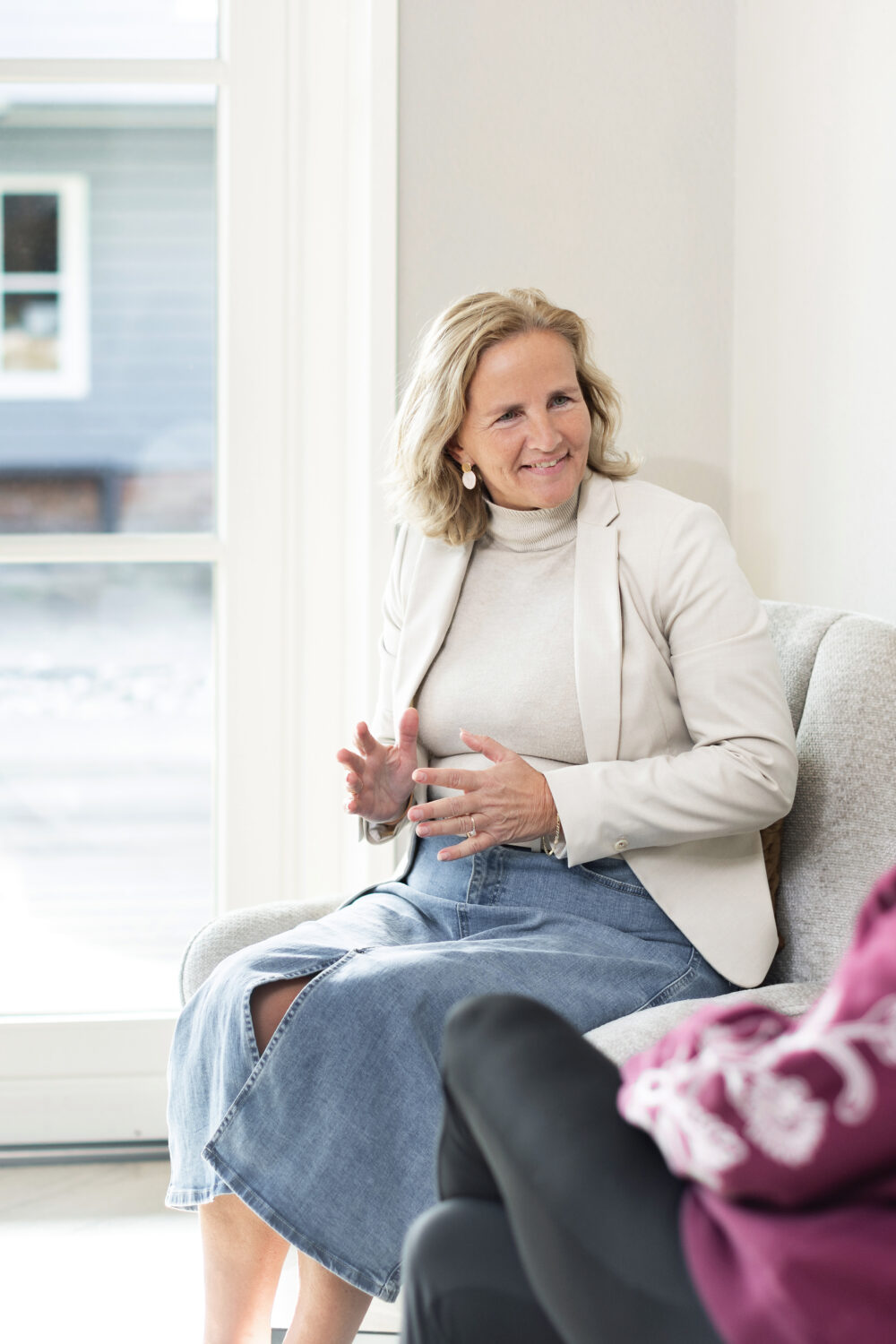 Vrouw in beige blazer zit in grijze fauteuil, kijkt naar een andere vrouw.
