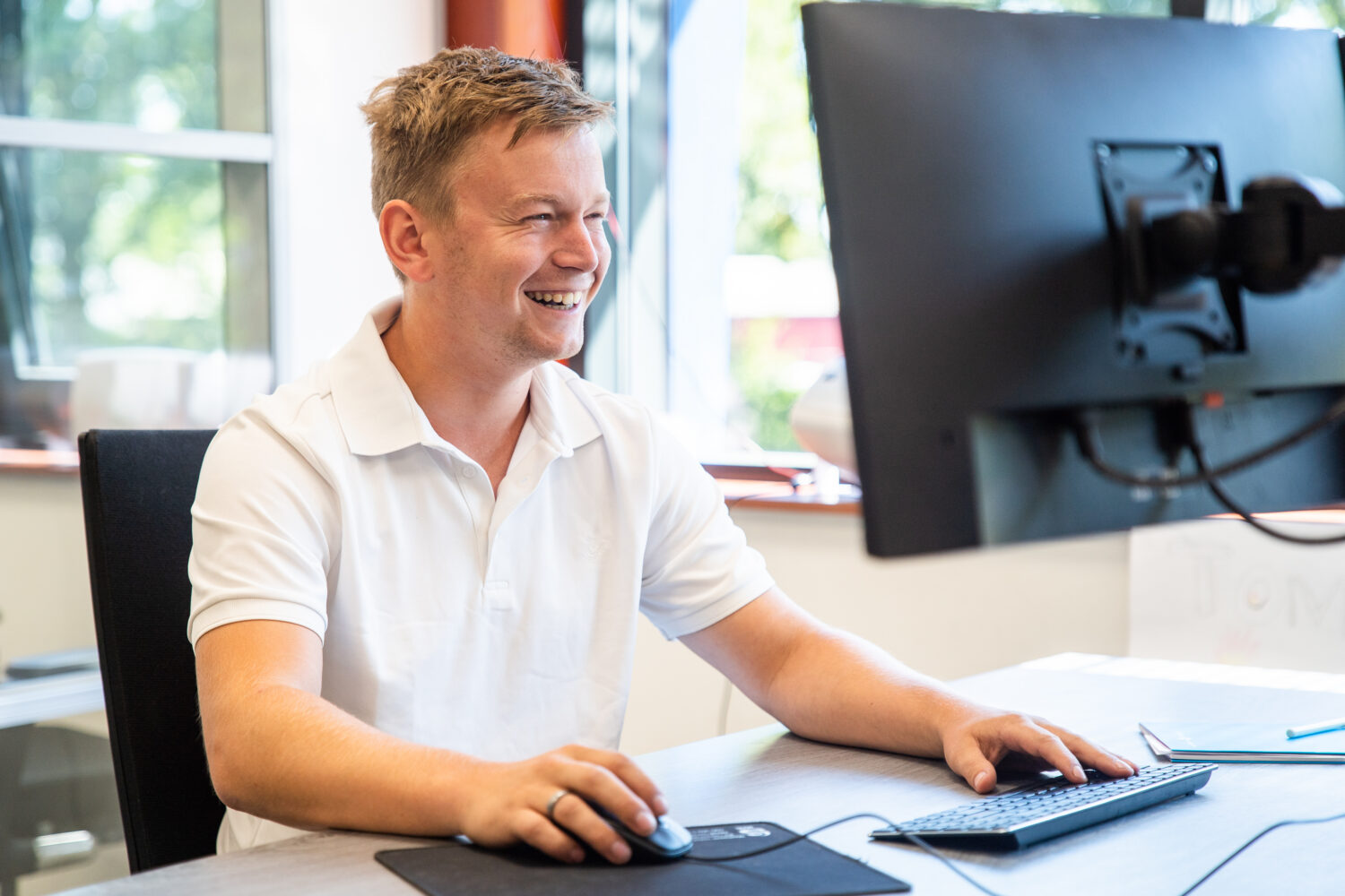Een lachende man zit achter een computerscherm met toetsenbord en muis.